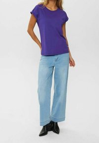 Violetti puuvillainen T-paita käärittyine hihoineen, yhdistetty vaaleansinisiin leveälahkeisiin farkkuihin ja mustiin nilkkureihin. Yksinkertainen, rento muotoilu.