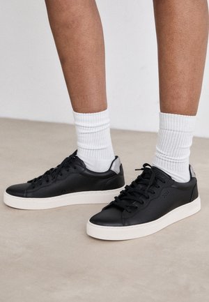 Czarne skórzane sneakersy z białymi podeszwami, o gładkiej fakturze, z sznurówkami i kontrastującym akcentem na tylnym obcasie, sparowane z białymi skarpetkami w prążki.