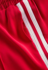 Sarkani sporta šorti, izgatavoti no gludas auduma, ar baltu sānu svītru, elastīgu jostasvietu un mīkstu tekstūru.