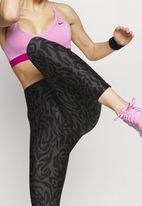 Donna in un reggiseno sportivo rosa e leggings neri a fantasia che solleva un ginocchio, indossando scarpe da ginnastica rosa e un braccialetto nero.