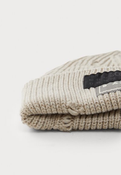 Bonnet en tricot beige avec un revers, présentant une étiquette noire sur le côté avec du texte blanc et des motifs géométriques. Texture douce.