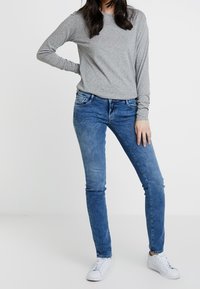 Šedé tričko s dlouhým rukávem, modré skinny džíny a bílé tenisky. Oblečení má ležérní design s měkkou texturou a čistými liniemi.