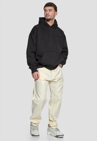 Svart oversized hettegenser med frontlomme, kombinert med off-white baggy bukser og grå sneakers med lyse detaljer. Glatt stoffstruktur.