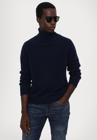 Marineblauer Rollkragenpullover aus weichem Strickstoff, kombiniert mit dunklen Jeans. Model trägt elegante schwarze Sonnenbrille. Schlichtes und modernes Erscheinungsbild.