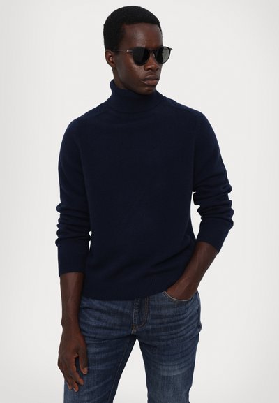Navy turtleneck sweater in soft knit fabric, paired with dark denim jeans. Model wearing sleek black sunglasses. Simple and modern appearance.