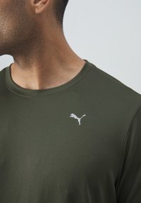 Camiseta de manga corta verde oliva con una tela texturizada, que presenta un logo blanco de Puma en el pecho y un diseño clásico de cuello redondo.