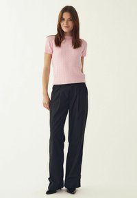 Roosa kootud lühikeste varrukatega kampsun tekstuurse mustriga, mida on kombineeritud mustade laiade pükstega ja läikivate mustade kingadega. Lihtne taust.