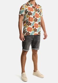 Hombre con una camisa de flores colorida de manga corta, pantalones cortos de mezclilla oscuros arremangados y zapatillas beige, posando frente a un fondo plano.