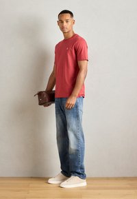 Camiseta de cuello redondo roja, de material de algodón, combinada con jeans claros desgastados. Sosteniendo un pequeño bolso de cuero marrón. Zapatillas blancas.
