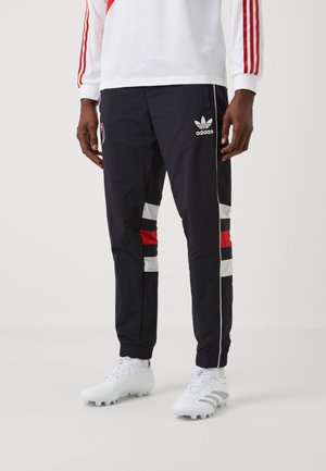 Person trägt schwarze Adidas-Trainingshose mit weißen und roten Streifen, weiße Sportschuhe und ein weißes Oberteil mit roten Streifen an den Ärmeln.