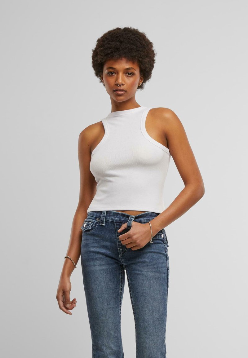 Top in cotone bianco a costine con collo alto, dalla vestibilità aderente e senza maniche. Abbinato a jeans in denim blu con taglio classico.
