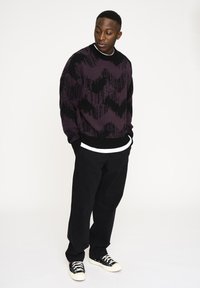 Schwarzer und lilafarbener gemusterter Strickpullover mit lockerer Passform, der ein Zickzackmuster aufweist, kombiniert mit schwarzen Hosen und schwarzen Turnschuhen.