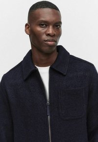 Merinovillasta valmistettu laivastonsininen takki, jossa on vetoketjukiinnitys, terävä kaulus ja yksi läpällinen tasku. Teksturoitu kangas, pehmeä pinta ja istuva malli.