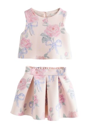 Niña joven con cabello rizado, vestida con un vestido blanco con estampados de flores rosas y lazos azules, y zapatos rosas tipo Mary Jane, de pie y mirando hacia un lado.