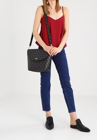 Bolso cruzado negro con estampado, tapa curva, herrajes en plata y correa ajustable, combinado con una blusa roja y jeans azules.