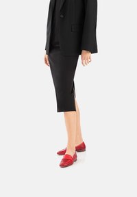 Scarpe slip-on rosse con un design perforato, dotate di una fascia sulla parte superiore e di un tacco basso, abbinate a una gonna nera e a un blazer.