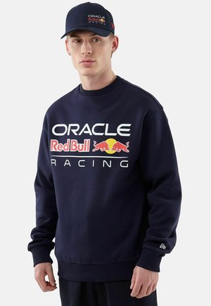 Młody mężczyzna ubrany w granatową bluzę Oracle Red Bull Racing i pasującą czapkę stoi na białym tle.