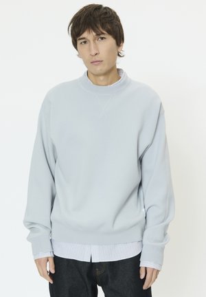 Felpa azzurro chiaro con scollo rotondo e polsini a coste, indossata sopra una camicia bianca a righe, con una vestibilità comoda.