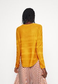 Senfgelbes, langärmeliges Strickoberteil mit strukturiertem Design, abgerundetem Saum und leichten seitlichen Schlitzen, kombiniert mit einem gemusterten Rock.