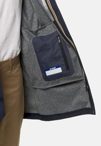 Giacca impermeabile navy con interni grigi. Include una tasca con zip e un'etichetta blu per informazioni personali. Texture liscia.