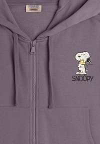 Haut à capuche zippé violet en tissu doux, avec un graphique de Snoopy et du texte. Comprend une poche avant et un capuchon avec cordon de serrage.