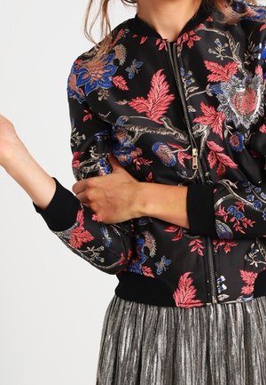 Femme portant une veste bomber noire à fleurs rouges et bleues et une jupe plissée métallique, les bras croisés et levés.