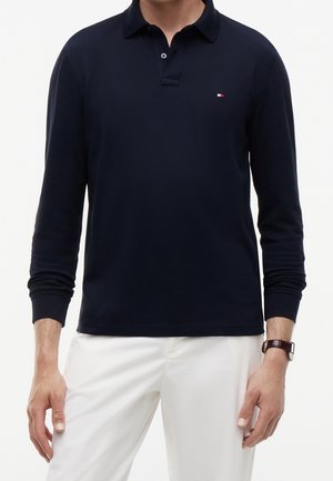 Homme portant un polo bleu marine à manches longues avec un petit logo sur la poitrine et un pantalon blanc, montre-bracelet visible au poignet gauche.