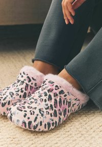 Leopardprint hjemmesko med pink pelsfor, lavet af blødt materiale, med ventilationshuller og en manchet-design; parret med mørkegrå bukser.