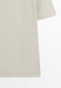 T-shirt beige à manches courtes avec ourlet doublement surpiqué visible et bord de manche sur fond blanc.