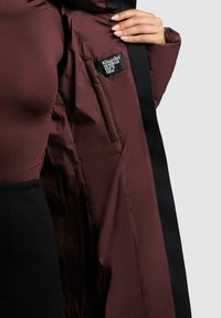 Eine kastanienrote Steppjacke mit glatter Außenseite und schwarzen Akzentstreifen, ausgestattet mit einer Innentasche, die mit einem Branding versehen ist.