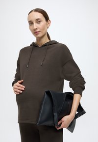 Καφέ βαμβακερό φούτερ με χαλαρή εφαρμογή και ρυθμιζόμενο κορδόνι, συνδυασμένο με μεγάλη μαύρη δερμάτινη τσάντα ώμου, που κρατεί ένα έγκυος άτομο.