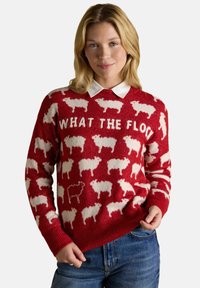 Frau trägt einen roten Pullover mit weißen Schafmustern und dem Text "WHAT THE FLOCK", kombiniert mit blauen Jeans und einem weißen Hemd darunter.