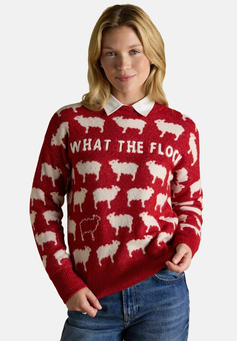 Frau trägt einen roten Pullover mit weißen Schafmustern und dem Text "WHAT THE FLOCK", kombiniert mit blauen Jeans und einem weißen Hemd darunter.