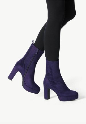 Jambes en leggings noirs portant des bottines compensées en daim violet avec talons carrés et fermetures éclair latérales sur fond blanc.