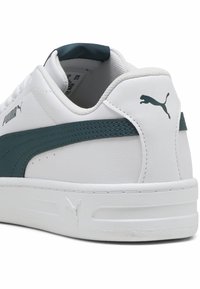 Vita sneakers med en slät finish, med en mörkt teal rand längs sidan, PUMA-logotyp och texturerade hälaccenter. Platt sula.