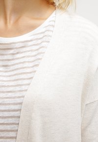 Ljusbeige cardigan lager över en vit randig T-shirt. Cardiganen har ett mjukt, texturerat stickat material med en öppen frontdesign.