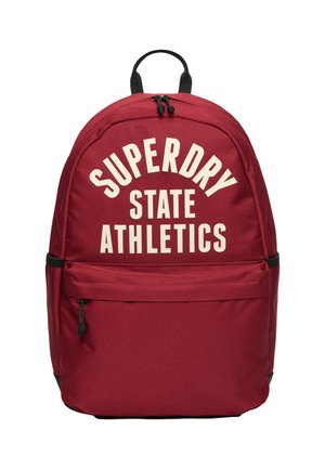 Mochila roja con asa y cremallera negra, bolsillo frontal y texto en blanco en negrita "SUPERDRY STATE ATHLETICS" en el compartimento principal.