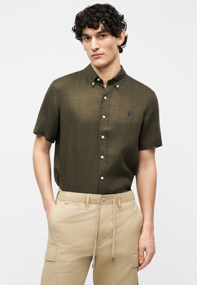 Άνδρας με σγουρά σκουρόχρωμα μαλλιά, που φοράει πουκάμισο ελιάς με κοντά μανίκια και μπεζ παντελόνι cargo, με τα χέρια στις τσέπες.