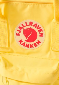 Dzeltena Fjällräven Kanken mugursoma ar apaļu logotipu, kurā attēlots sarkans lapsa, ar izsmalcinātu šuvumu un teksturētiem plecu siksnām.