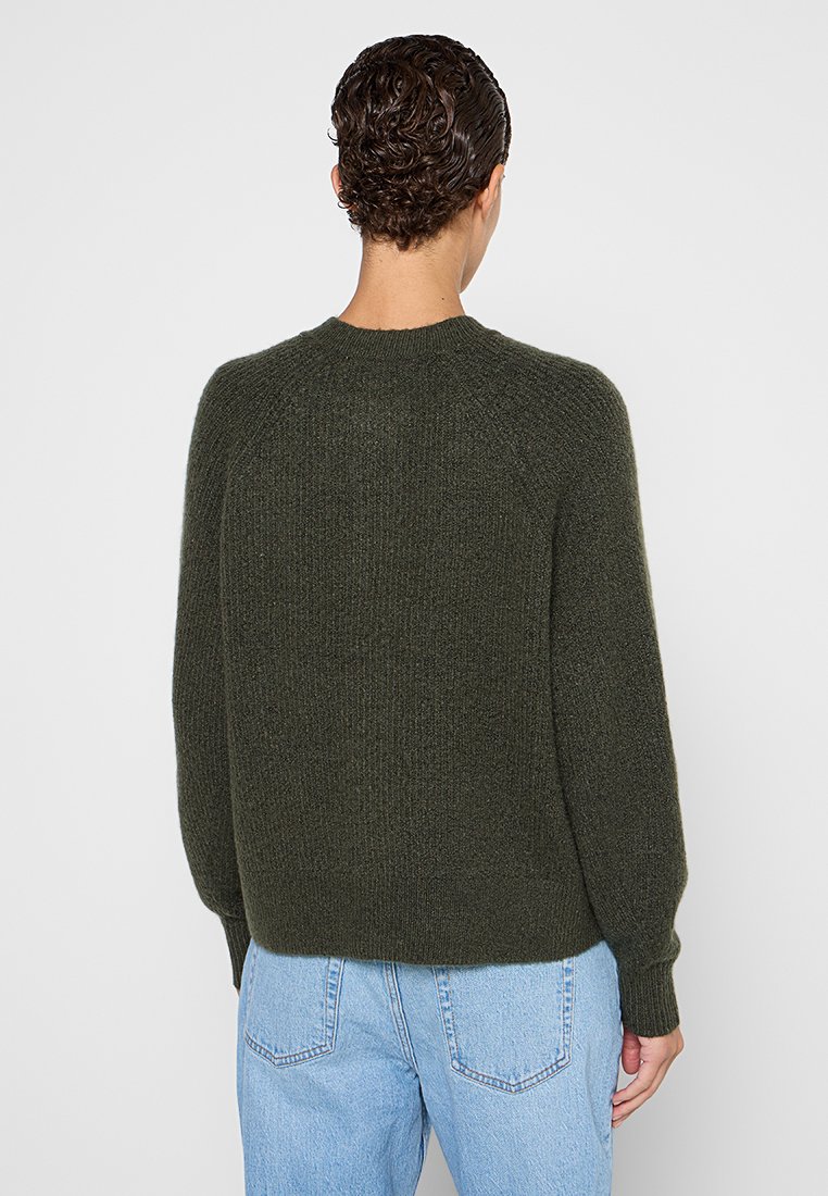 Dunkelgrüner gestrickter Pullover mit rundem Halsausschnitt, Raglanärmel und gerippten Bündchen. Weiche Textur und lockere Passform. Getragen mit hellblauen Jeans.