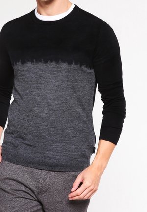 Hombre con un suéter de manga larga y cuello redondo, con la sección superior negra y la sección inferior gris texturizada, combinado con pantalones grises.