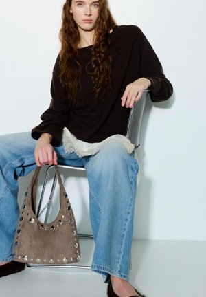 Mlada ženska z dolgimi valovitimi lasmi sedi na kovanskem stolu, nosi črno pulover, svetlo modre kavbojke in drži rjavo semiš torbico z zakovicami.