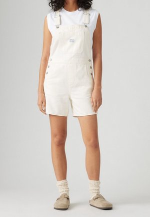 Białe bawełniane ogrodniczki z regulowanymi ramiączkami, kieszenią z przodu i bocznymi guzikami. Noszone na białą bezrękawnik i połączone z beżowymi butami.