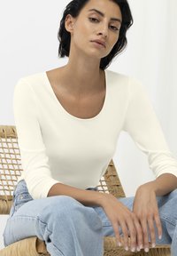 Biała bluzka z długim rękawem i okrągłym dekoltem, wykonana z miękkiego materiału. Modelka nosi jasnoniebieskie dżinsy, siedząc na plecionej krześle. Neutralne tło.