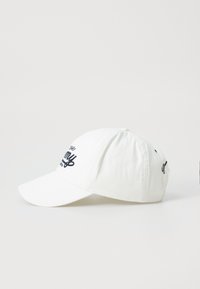 Casquette de baseball en coton blanc avec une visière courbée, comportant un texte brodé et des accents de logo en bleu marine sur l'avant et sur le côté.