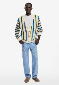 Cardigan a maglia multicolore con righe verticali sopra una T-shirt bianca, abbinato a jeans azzurri chiari con motivi astratti colorati e scarpe marroni.