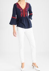 Blusa blu navy con ricami floreali rossi, scollatura a V e maniche a campana, abbinata a pantaloni slim-fit bianchi e tacchi bianchi a punta.