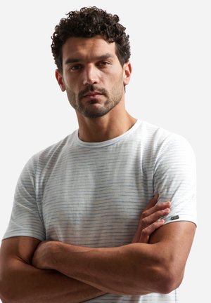 Man met krullend donker haar en baard, gekleed in een wit en lichtblauw gestreept T-shirt, met gekruiste armen, kijkt recht vooruit.