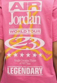 Ροζ πουκάμισο με μεγάλα λευκά γράμματα "Air Jordan", "World Tour", αριθμός 23, κίτρινη σιλουέτα καλαθοσφαιριστή και τη λέξη "Legendary" στο κάτω μέρος.
