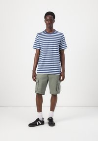 Blauw-wit gestreept T-shirt gecombineerd met olijfgroene cargo shorts. Model draagt zwarte sneakers met witte accenten en witte sokken.